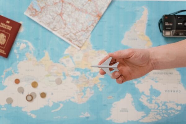 Hand holding a toy airplane over a world map, surrounded by coins, camera, passport, travel insurance with pre-existing health conditions
