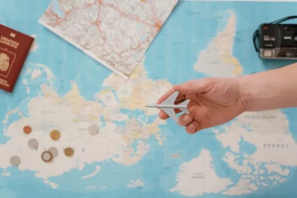 Hand holding a toy airplane over a world map, surrounded by coins, camera, passport, travel insurance with pre-existing health conditions