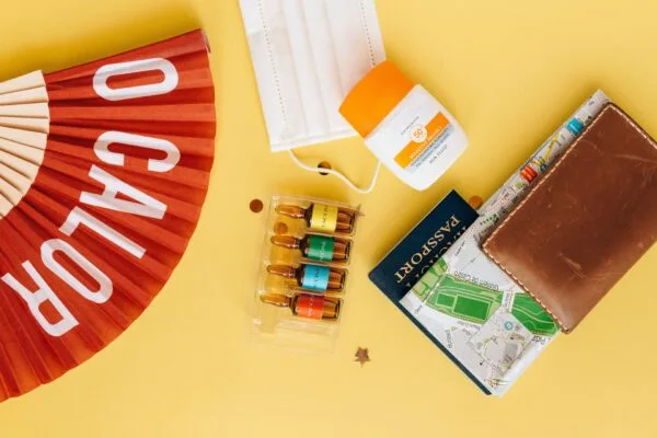 Flatlay of travel essentials like mask, vaccine, and sunscreen on yellow background