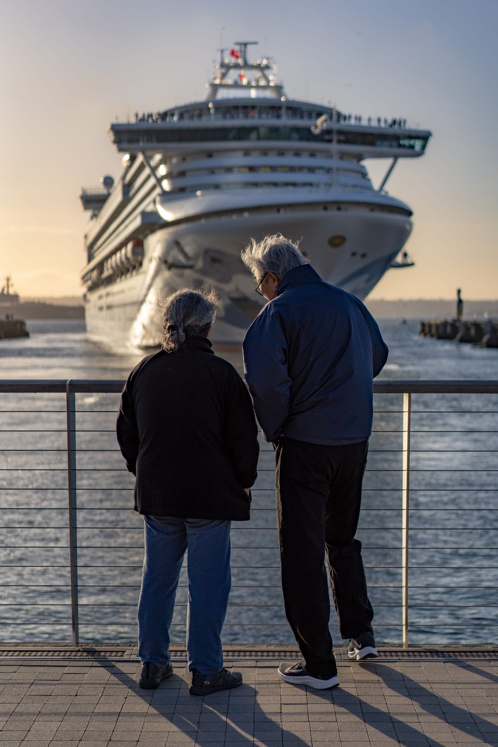 Cruising With Epilepsy: A Complete Guide, Senior couple standing at railing, admiring large cruise ship approaching dock at sunset.
