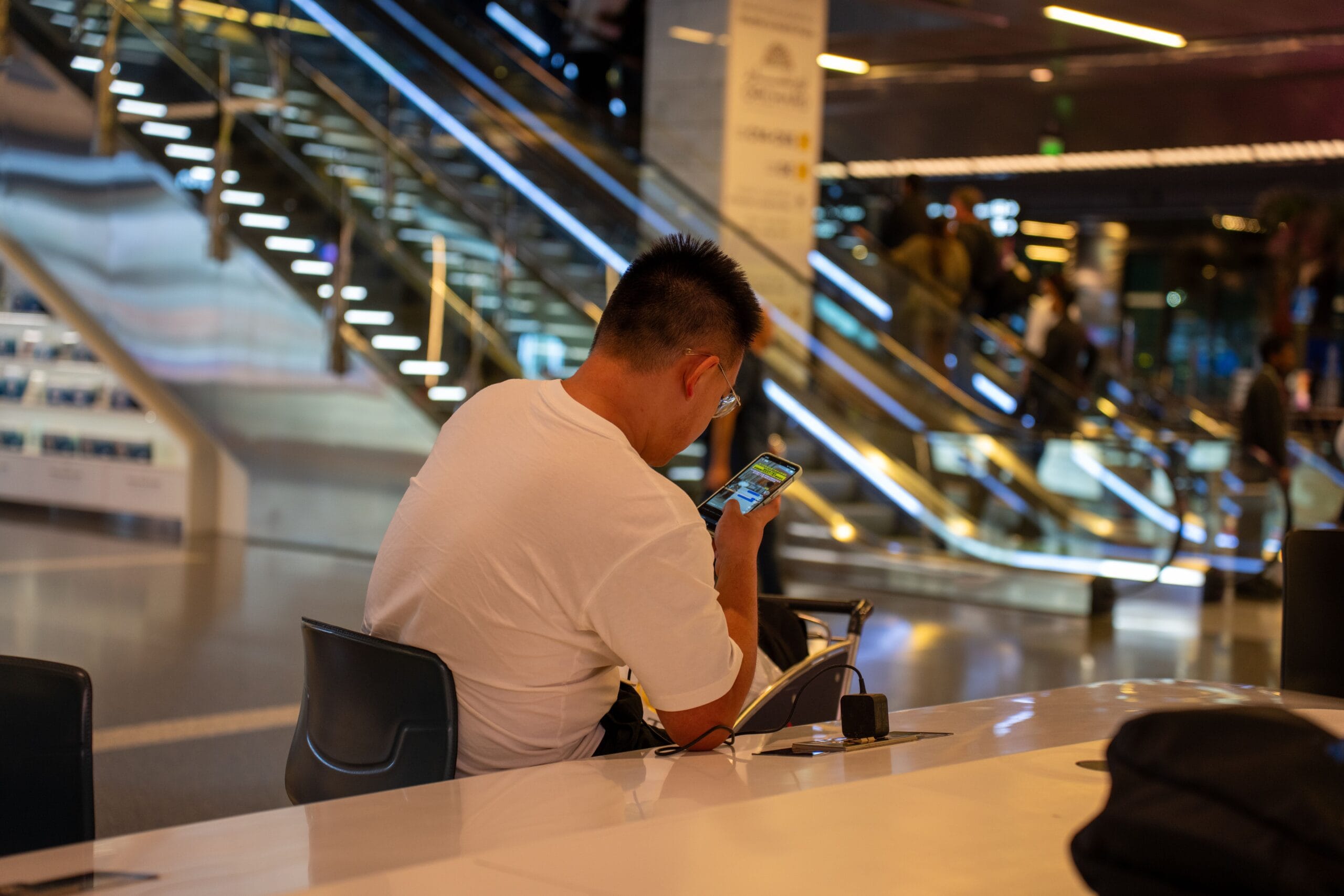 Flying After Hip or Knee Replacement: A Complete Guide, Adult using smartphone in modern airport terminal, Doha, Qatar. Busy travel scene with escalator.
