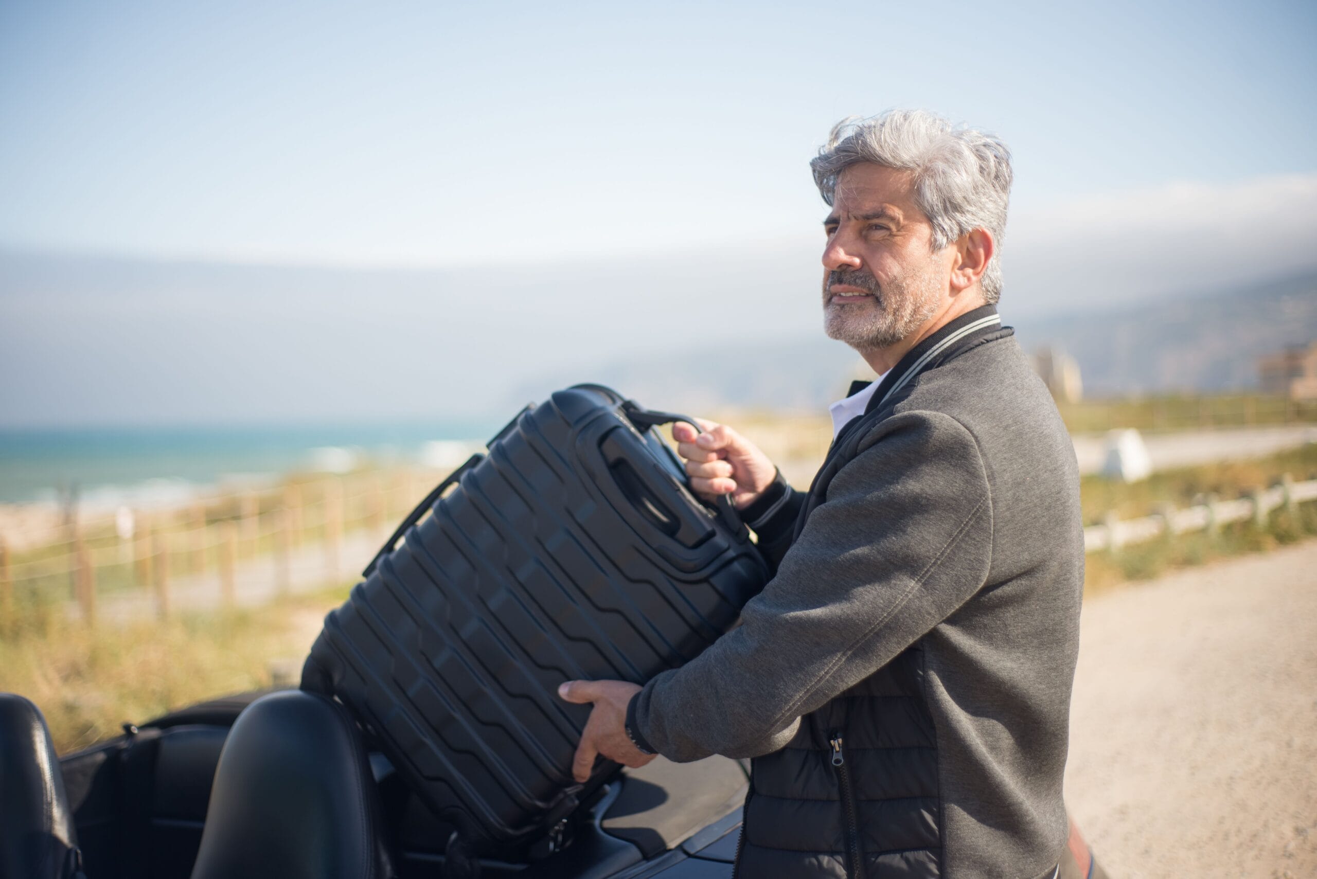 Travel insurance with a pacemaker or ICD - Senior travller man with suitcase by a convertible on a scenic coastal journey in Portugal.