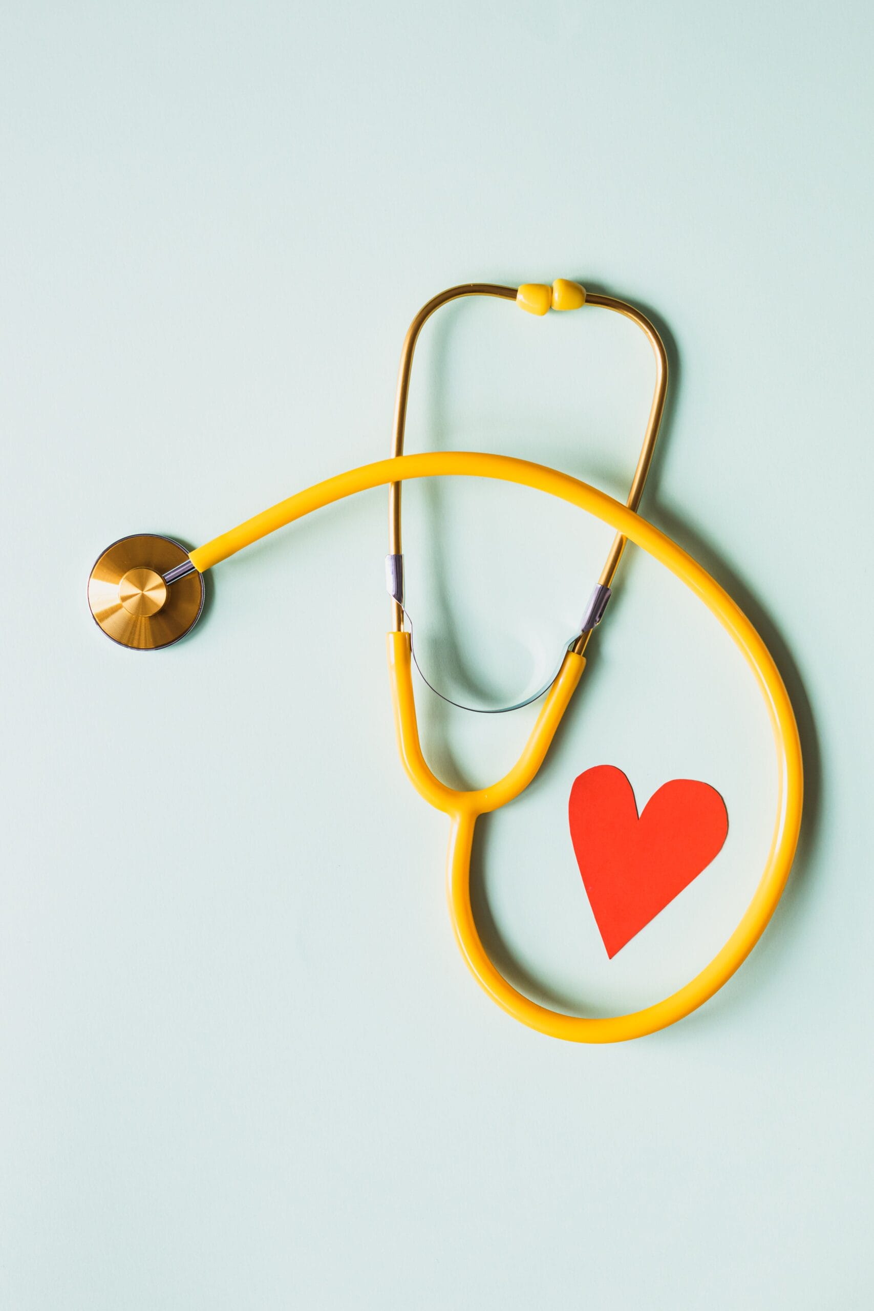 Travel Insurance With Heart Conditions, Yellow stethoscope arranged with a red heart cutout on a light green background, symbolizing heart health.